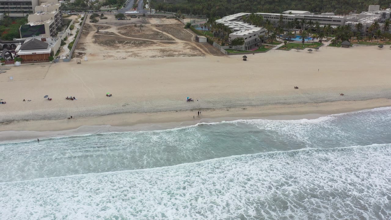 Aerial beach view with white surf, open sand, and buildings behind it.