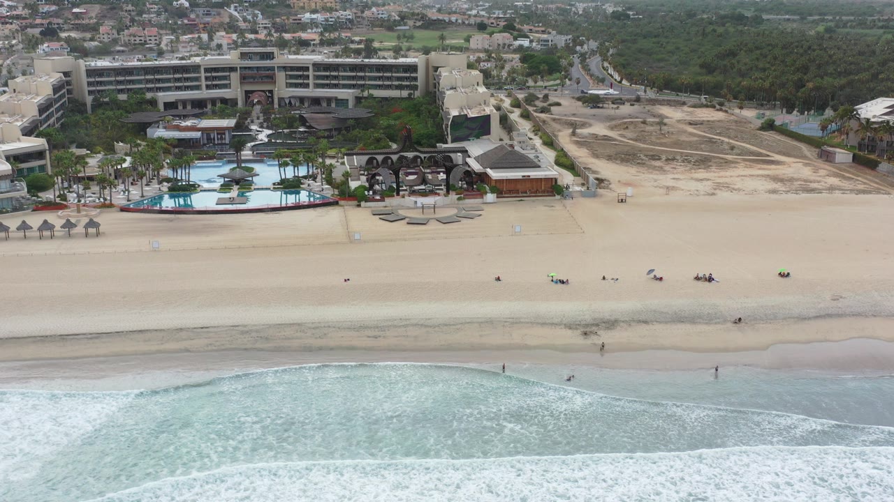 Aerial view of beach, surf, resort buildings, pools, roads, and inland greenery.