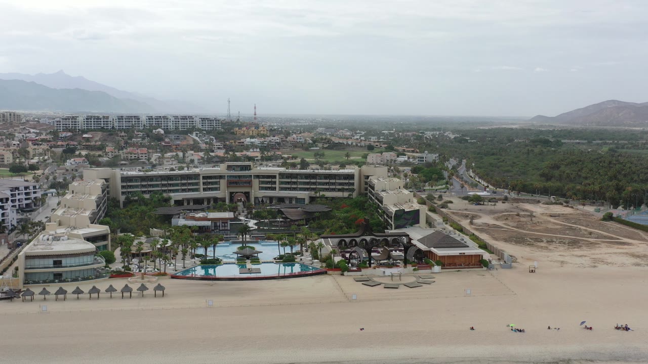 Aerial view in Los Cabos, Mexico, showing a wide sandy beach in front of resort buildings, pools, roads, green land, hazy hills, and no visible people.