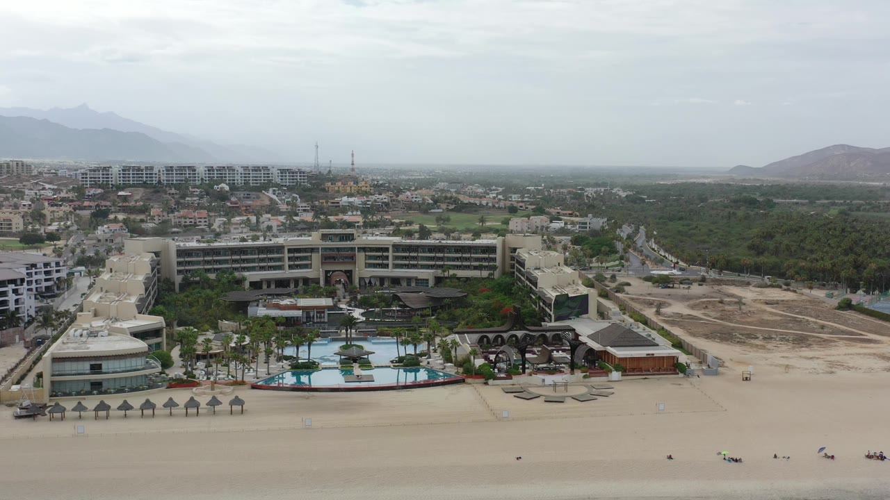 Beachfront resort complex with pools, palapas, roads, and green land beyond.