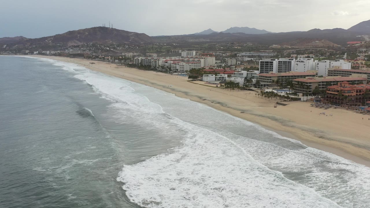 Aerial view in Los Cabos, Mexico showing gray-green surf breaking toward a broad sandy beach, beachfront buildings, brown hills, distant mountains, and no visible people.