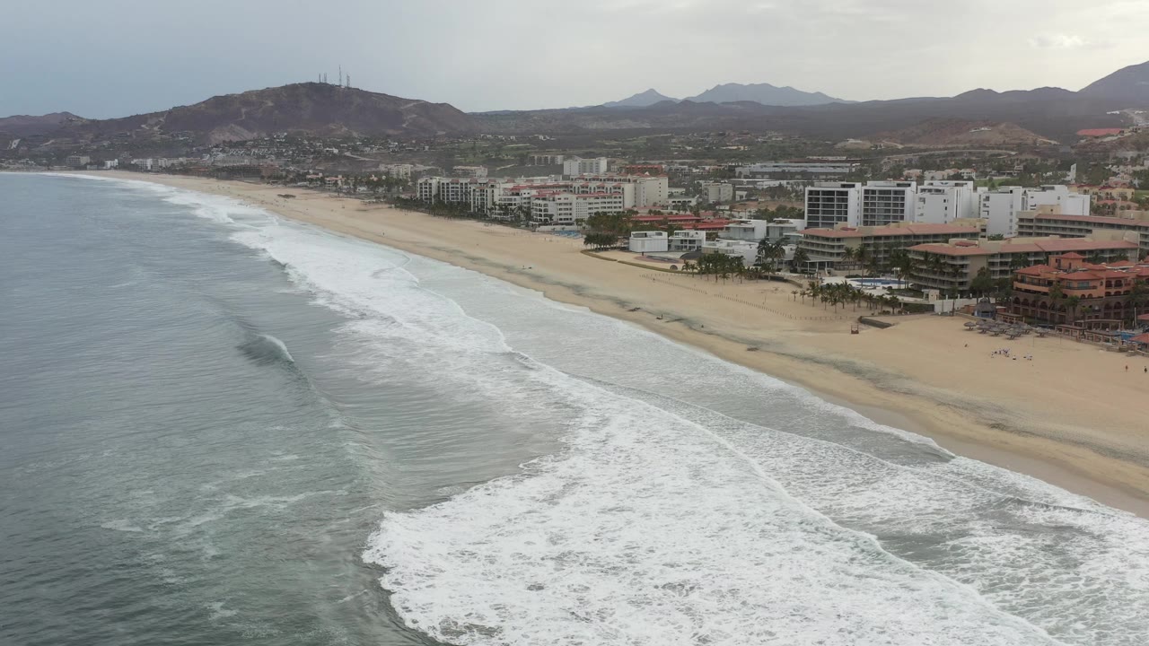 Aerial Los Cabos, Mexico frame with a large white wave band spreading over shallow water, a pale beach, modern buildings near the sand, hills beyond, and no visible people.