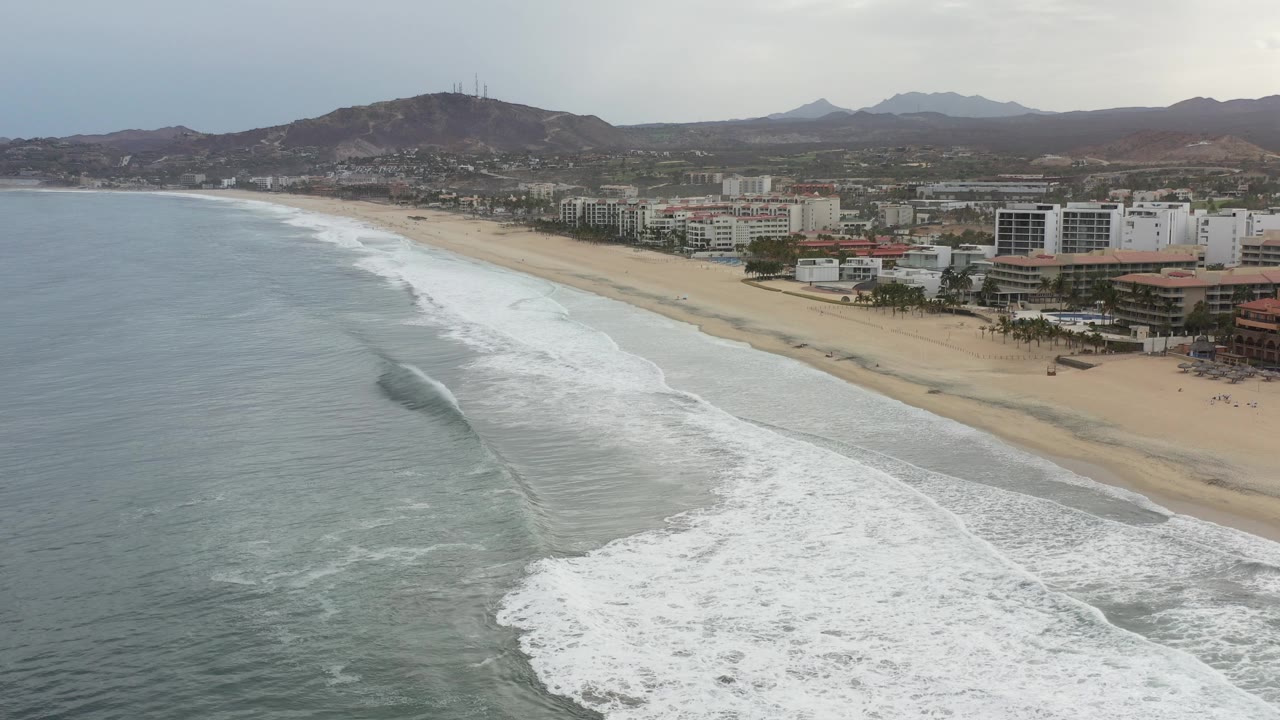 Diagonal aerial coastline view with surf, beach, buildings, and mountains under clouds.