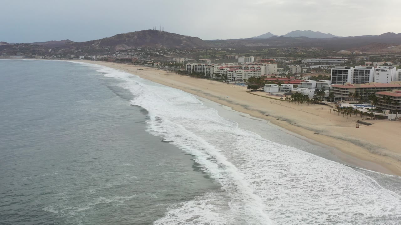 Elevated view over the Los Cabos shoreline with gray-blue water, white surf, sandy beach, hotel buildings, hills, and no visible people.