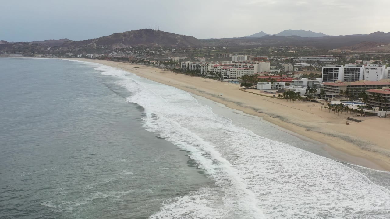 Los Cabos beach seen from above as parallel lines of foam advance toward sand, with hotel blocks and brown hills under a gray sky, no visible people.