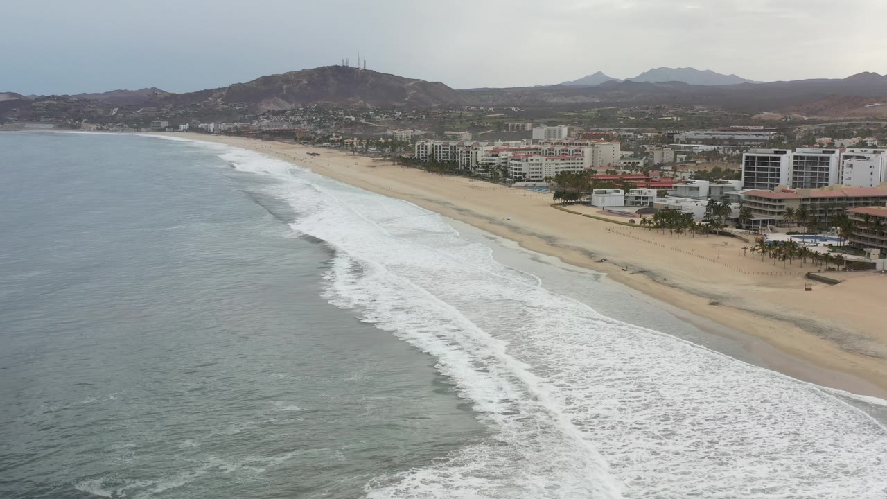 Wide coastline with surf, sand, hotels, and mountains under cloudy light.