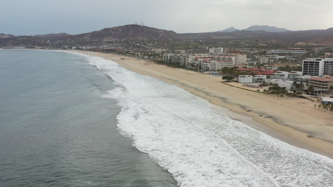 Aerial view of surf rolling onto a sandy beach beside a seaside town.
