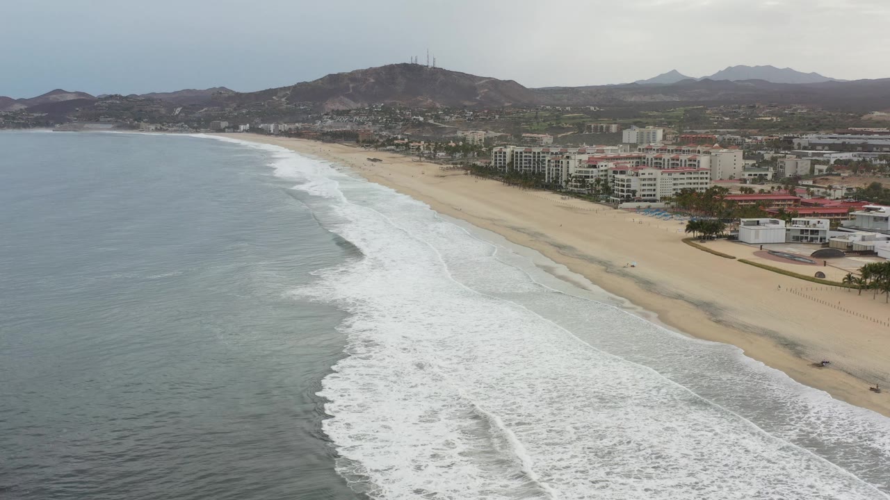 Los Cabos shoreline from above with a long white surf line curving beside tan sand, clustered low-rise buildings near the beach, hills in the distance, and no clearly visible people.