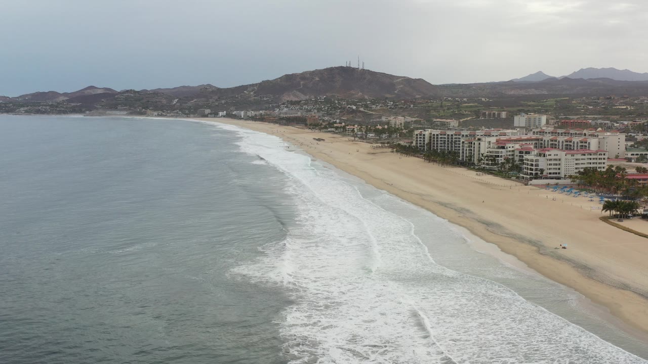 Wide aerial frame of the Los Cabos coastline with pale water at left, white surf bands reaching the sand, beachfront buildings to the right, mountains beyond, and no visible people.