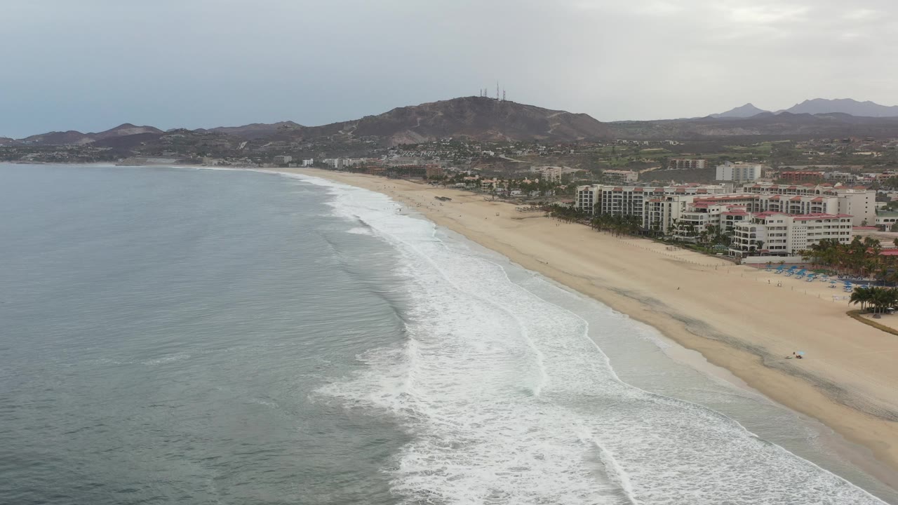 Long sandy shoreline with foamy waves and a mountain ridge above the city.
