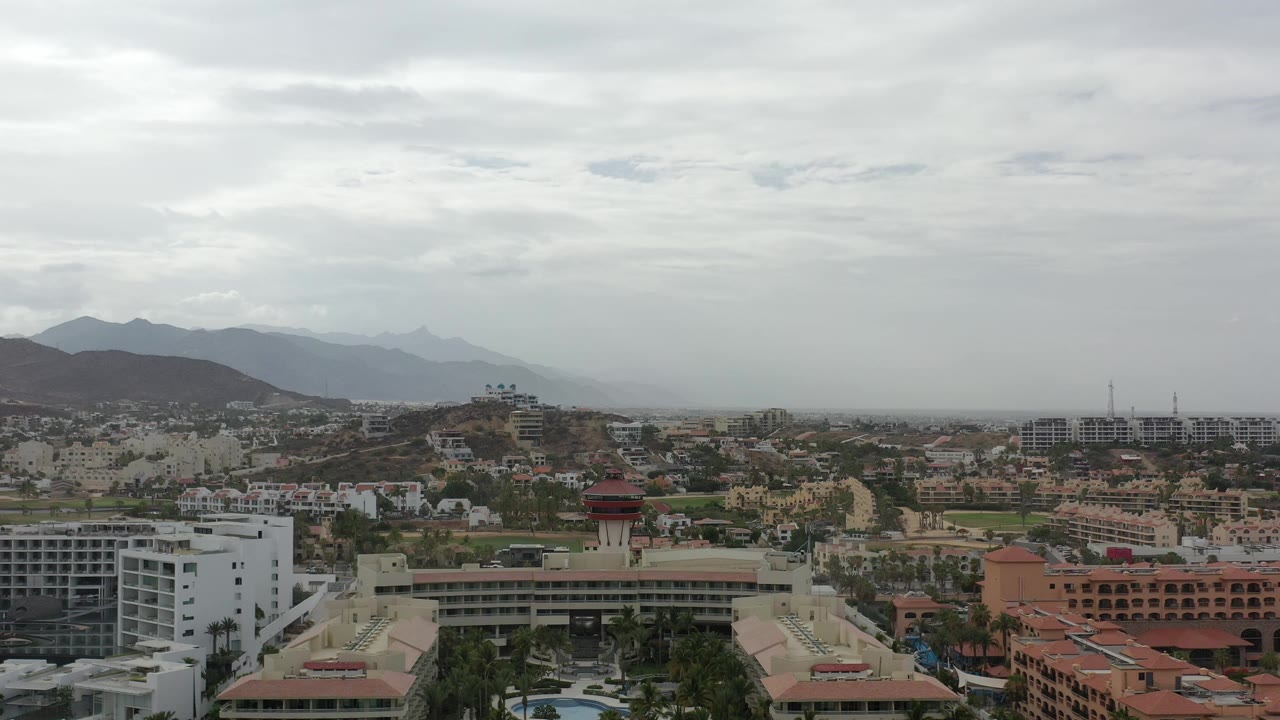 Wide elevated view over Los Cabos buildings and distant mountains under gray clouds.