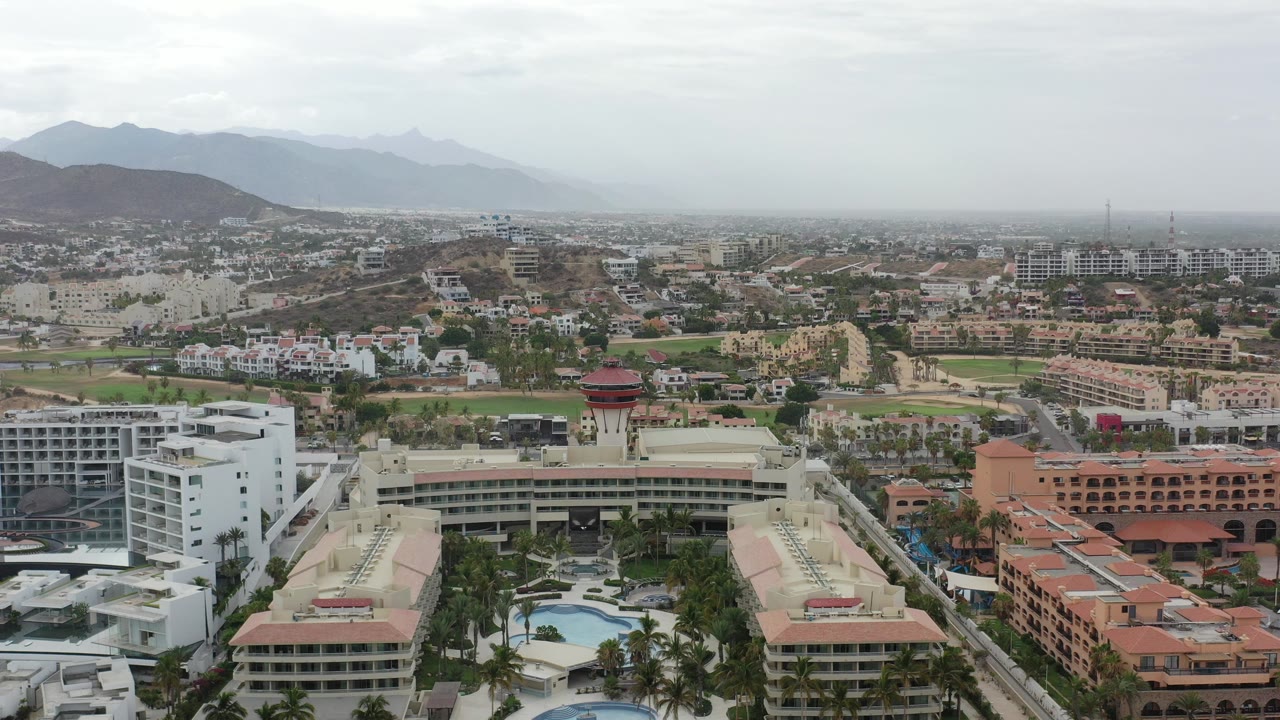 Aerial view of resort buildings, pools, city blocks, and mountains under a cloudy sky.