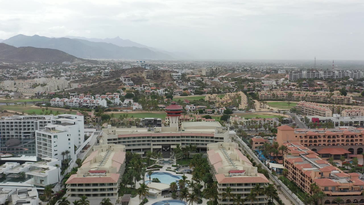 Los Cabos aerial view with the red-topped resort tower near the lower center, white hotel blocks and pool courtyards nearby, city streets behind, and no visible people.