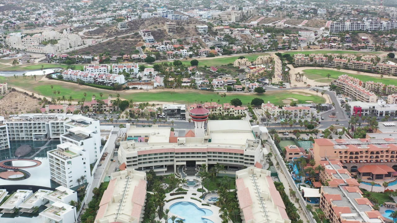 Aerial view over large resort buildings, pools, roads, green grounds, and hillside housing.