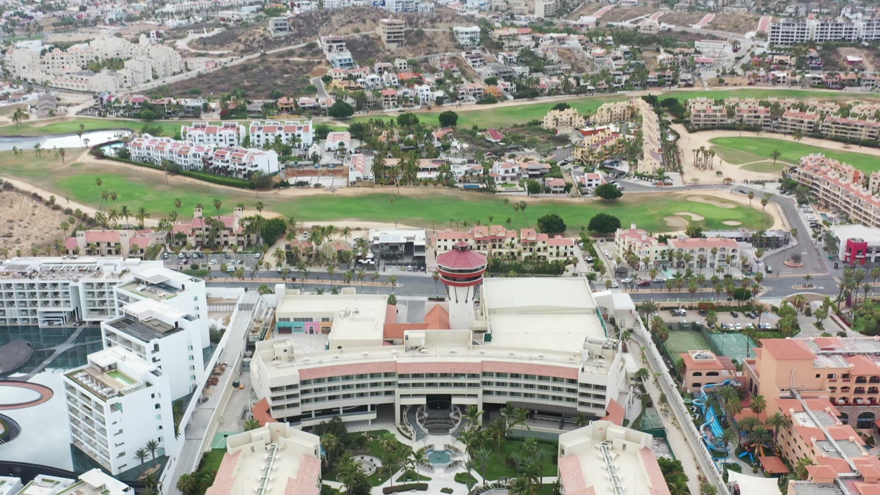Aerial view in Los Cabos showing the resort roofline closer to center, with pools, palm-lined roads, nearby villas, golf course, and no visible people.