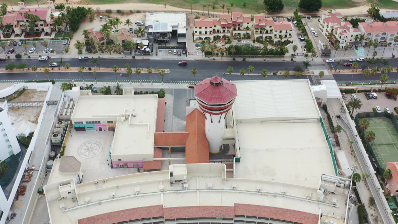 Los Cabos overhead frame with the tower slightly right of center, flat cream roofs, terracotta roof bands, a road, parked cars, and no visible people.