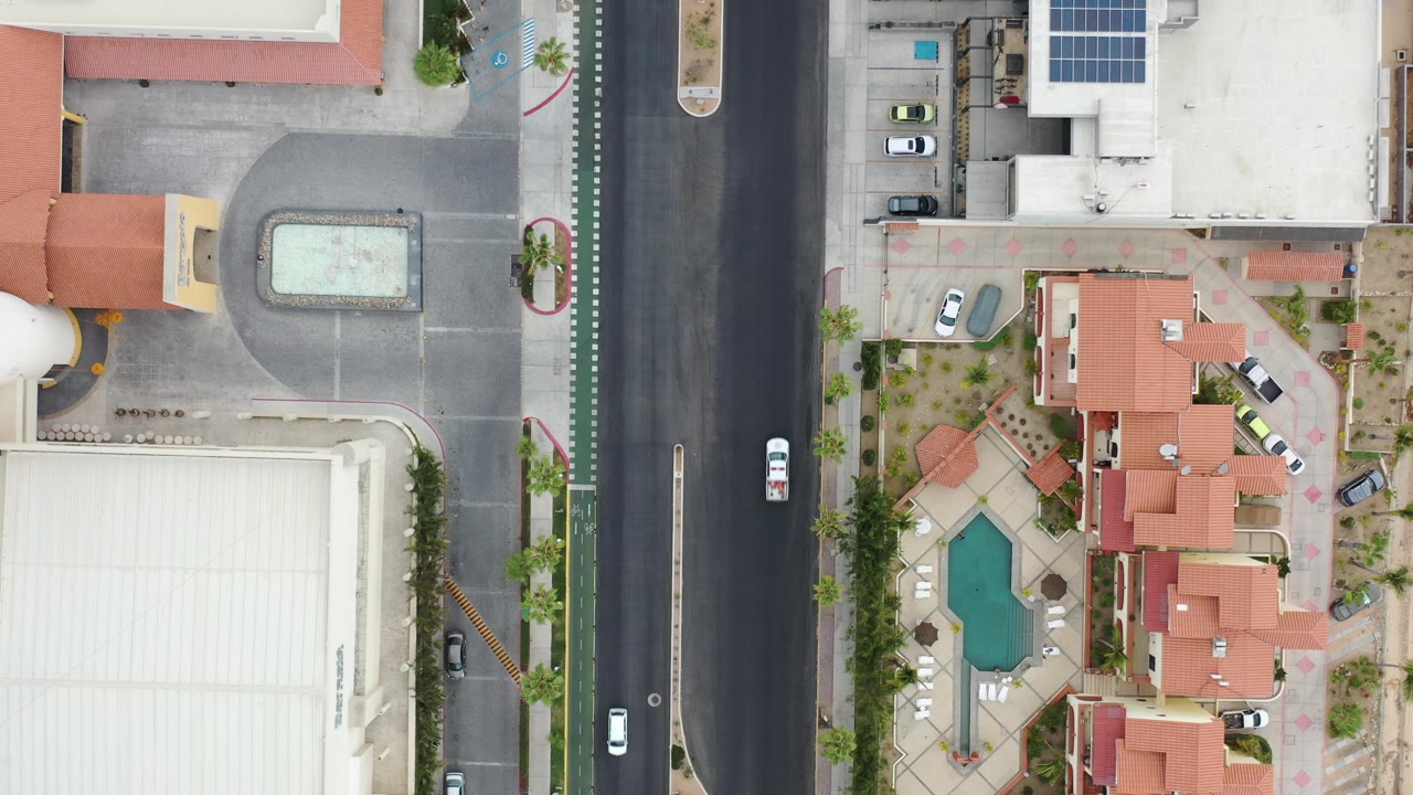 Overhead view of a white car on a residential street beside homes and a pool courtyard.