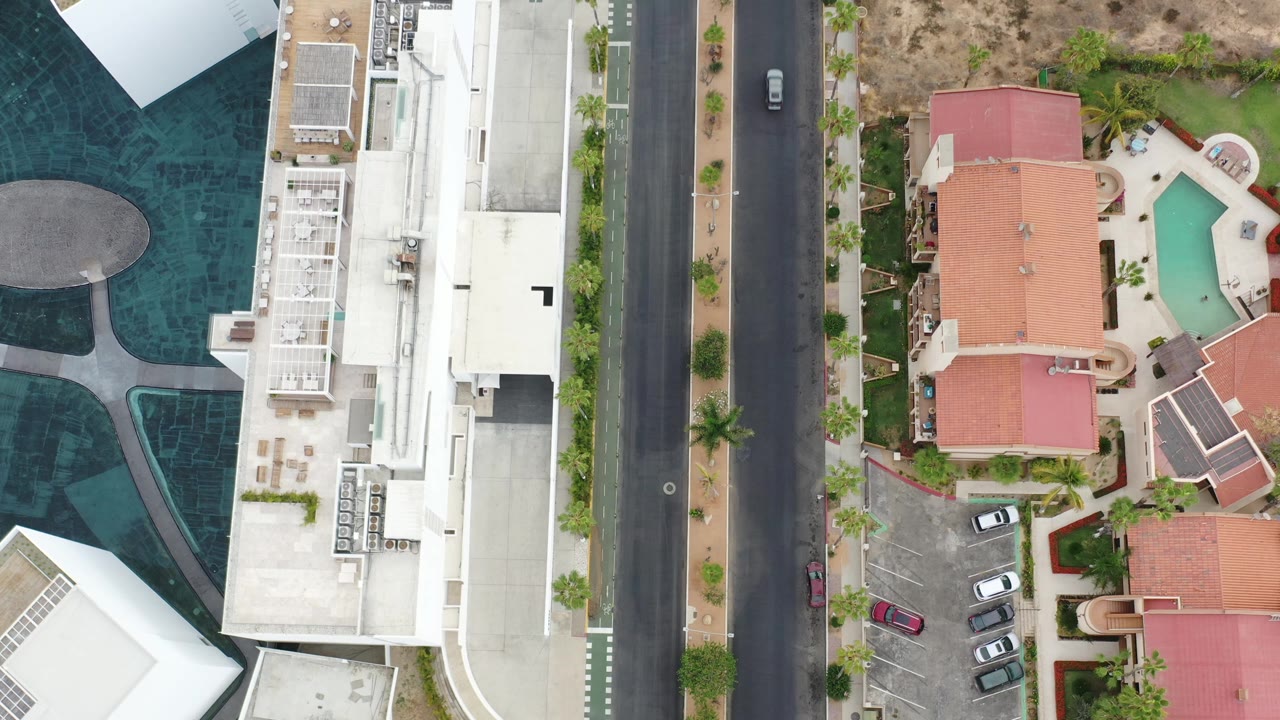 Los Cabos aerial view with the road still centered, a small dark car near the top of the street, white building roofs on the left, red-roofed residences on the right, pools, parked cars, and no visible people.