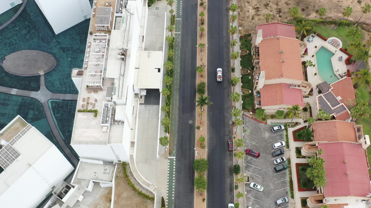 Overhead view of road, white buildings, red-roofed homes, pools, and parked cars.
