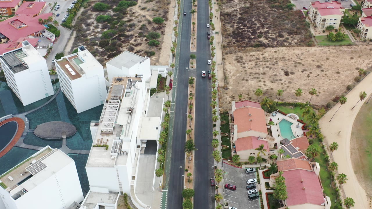 High aerial frame over Los Cabos showing white modern buildings, a straight road with cars, red-roof homes, dry lots, landscaping, and no visible people.