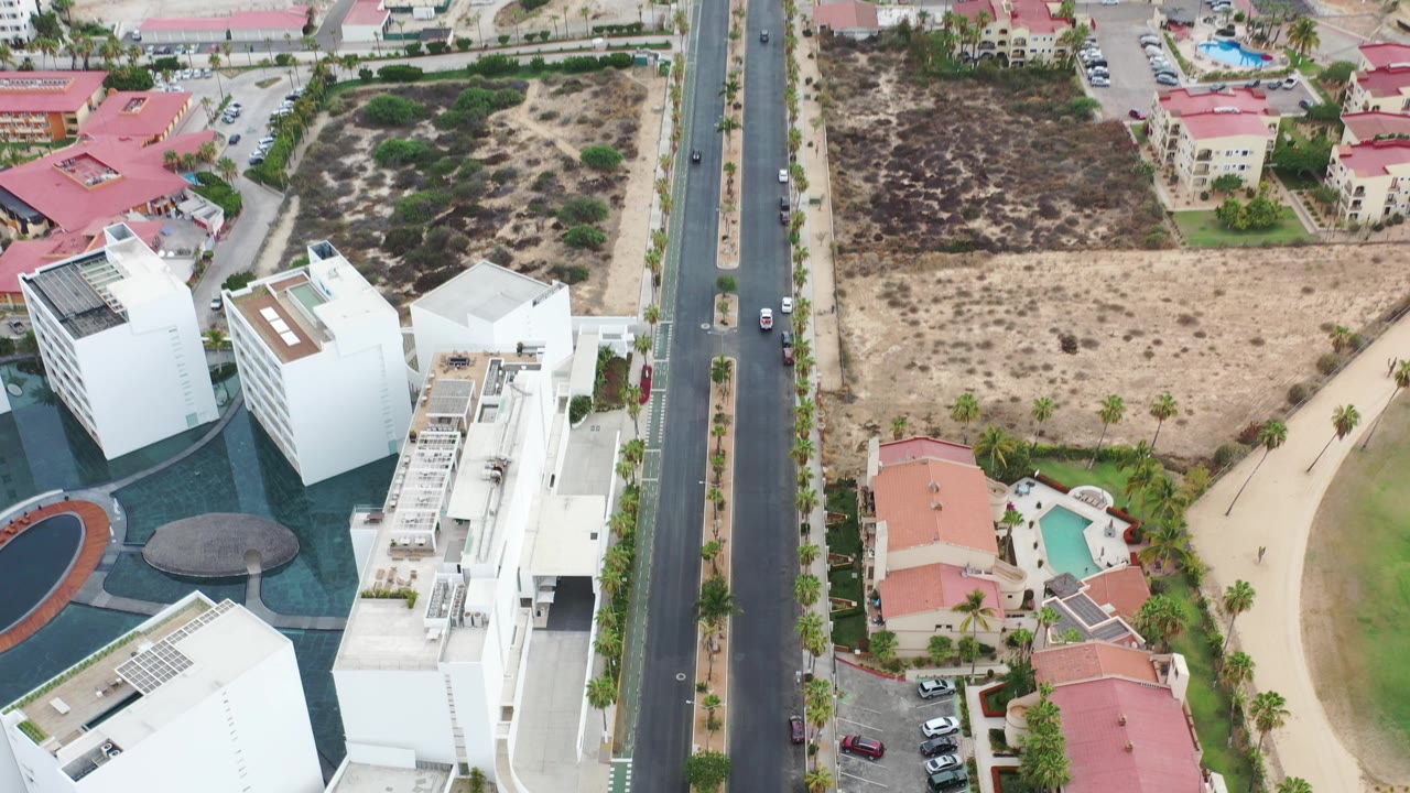 Los Cabos aerial frame showing white building rooftops, a vertical road with cars, red-roof villas, dry parcels, green landscaping, and no visible people.