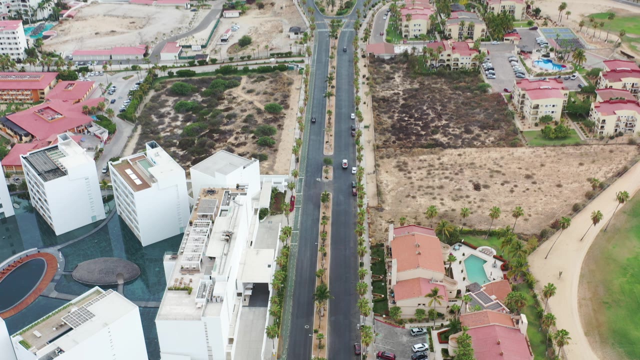 Wider aerial with road, resort-style buildings, homes, open land, beach, and ocean.