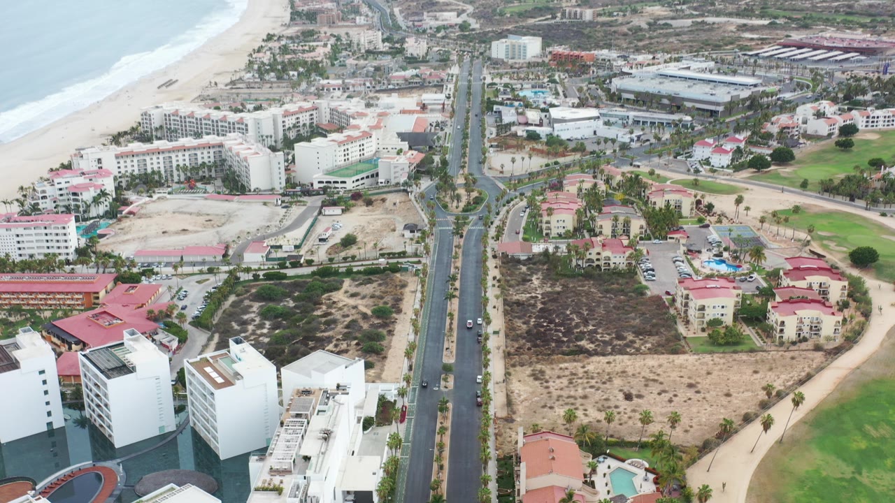 Opening aerial frame over Los Cabos, Mexico with the beach and surf at left, a straight road through beachfront buildings, dry lots, and distant mountains, with no visible people.