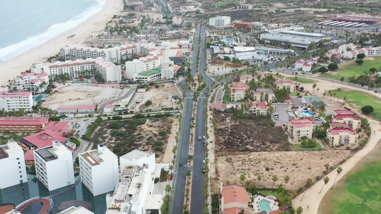 Aerial view in Los Cabos, Mexico with the road centered more tightly between beachfront buildings, sandy beach to the left, mixed rooftops below, and no visible people.