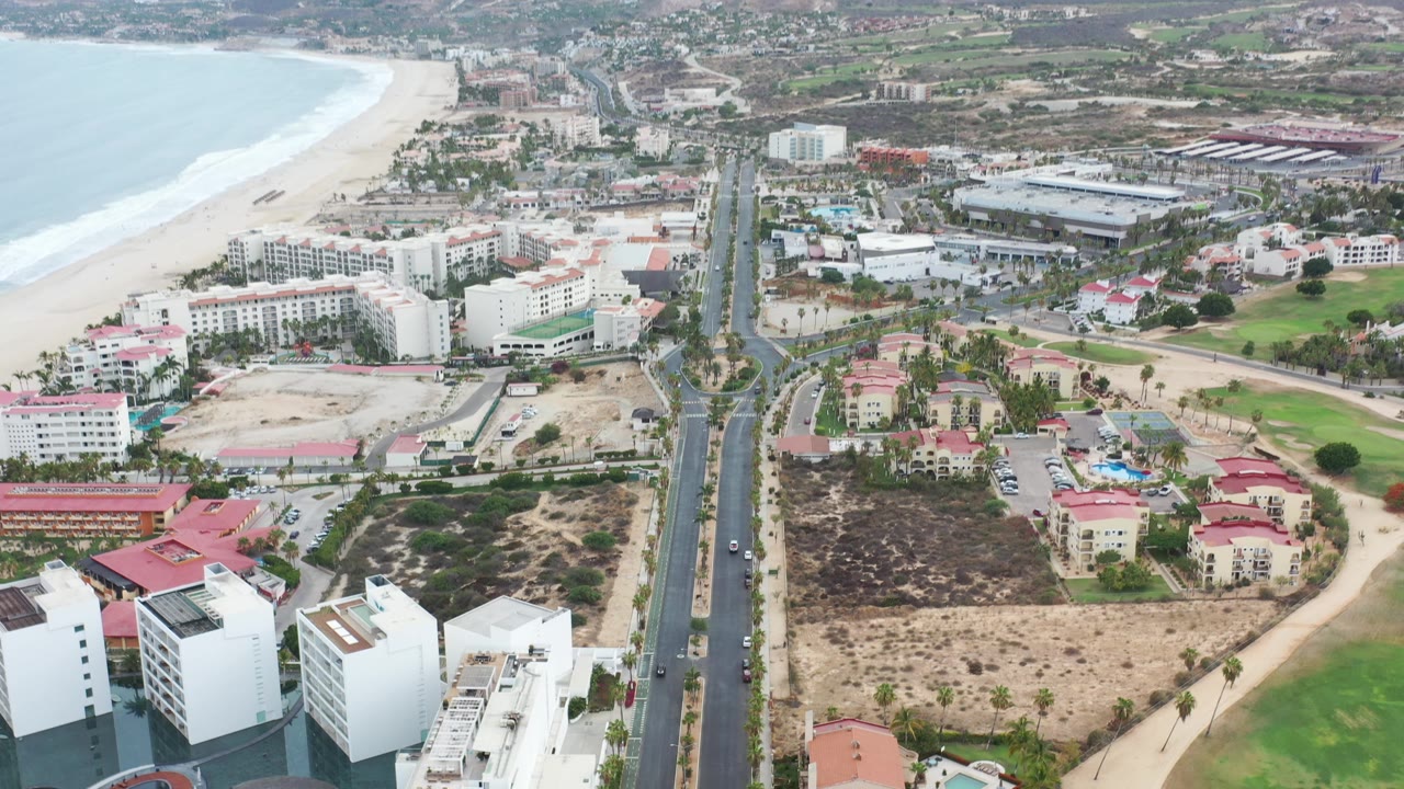 Coastal city aerial with ocean, beach, road, rooftops, and mountains.
