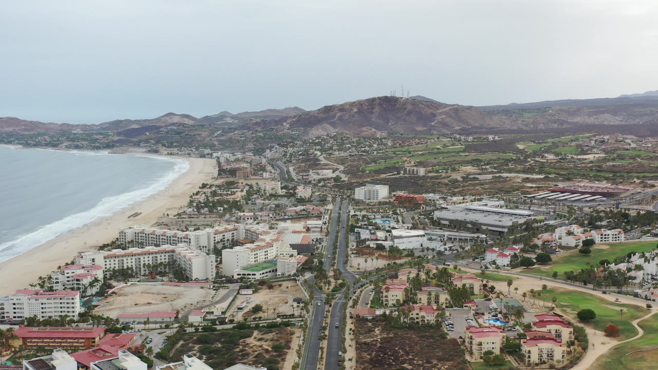 Aerial view over Los Cabos, Mexico, with the beach and surf at left, a main road running inland through buildings, mountains on the horizon, and no visible people.