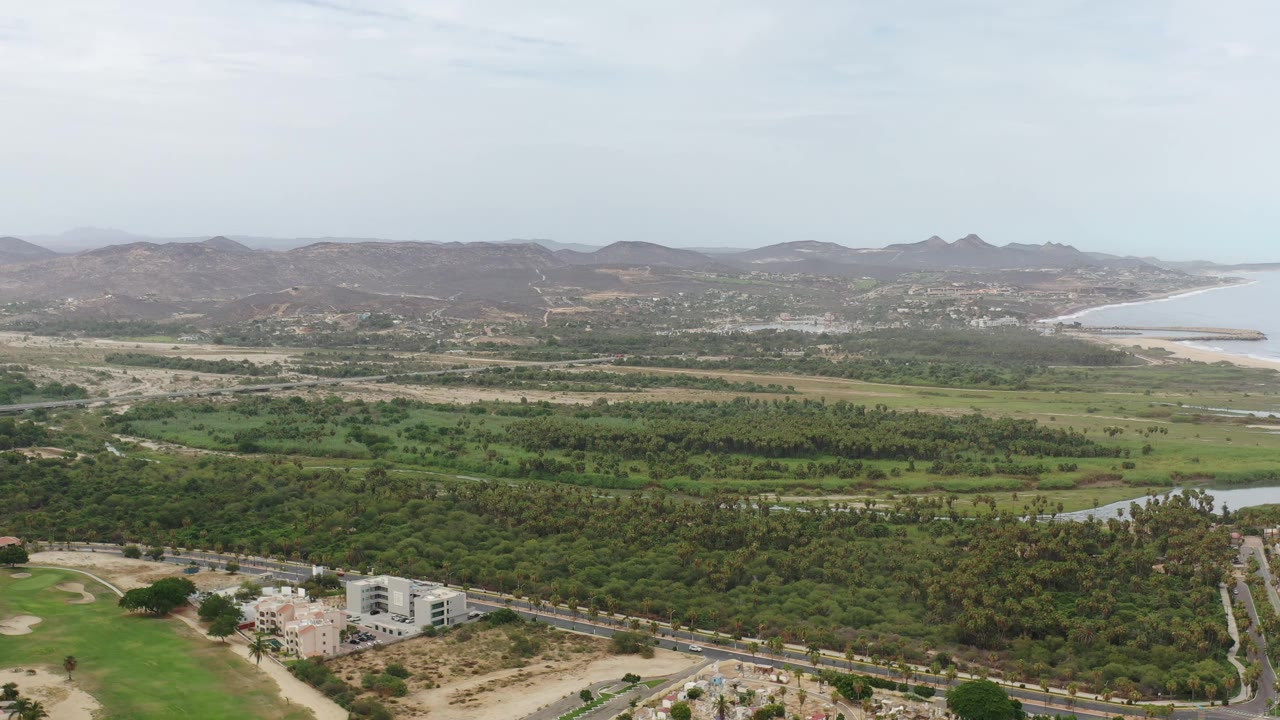 Aerial frame from Los Cabos showing the road and white buildings lower in frame, a broad green belt across the center, brown hills beyond, coastline at right, and no visible people.