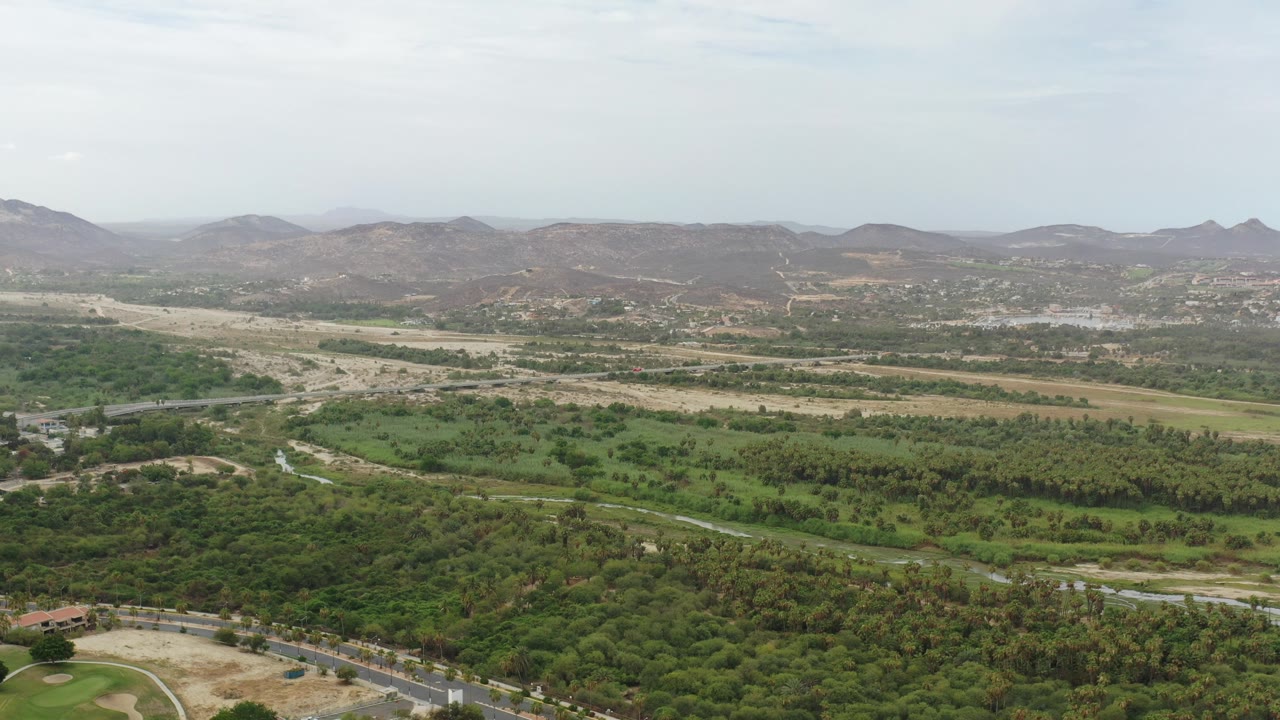 Wide aerial view over Los Cabos, Mexico, with green tree cover, winding roads, dry hills in the distance, pale sky, and no visible people.