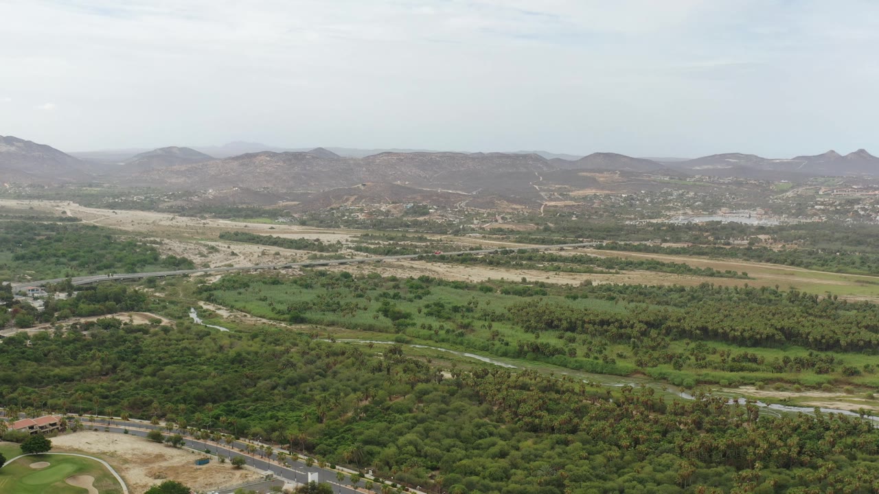 A broad Los Cabos aerial landscape shows dense green vegetation in the foreground, tan open ground, distant brown hills, and no visible people.