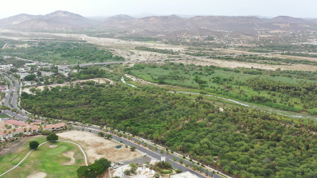 Aerial frame from Los Cabos, Mexico, with a road running through green vegetation, small buildings near the lower edge, pale dry wash areas beyond, and no visible people.