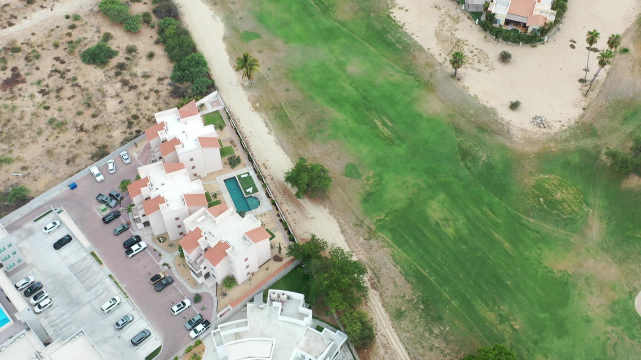 Overhead Los Cabos frame showing white residential buildings, parked cars, a small pool, sandy roads, desert scrub, and green landscaped ground with no visible people.