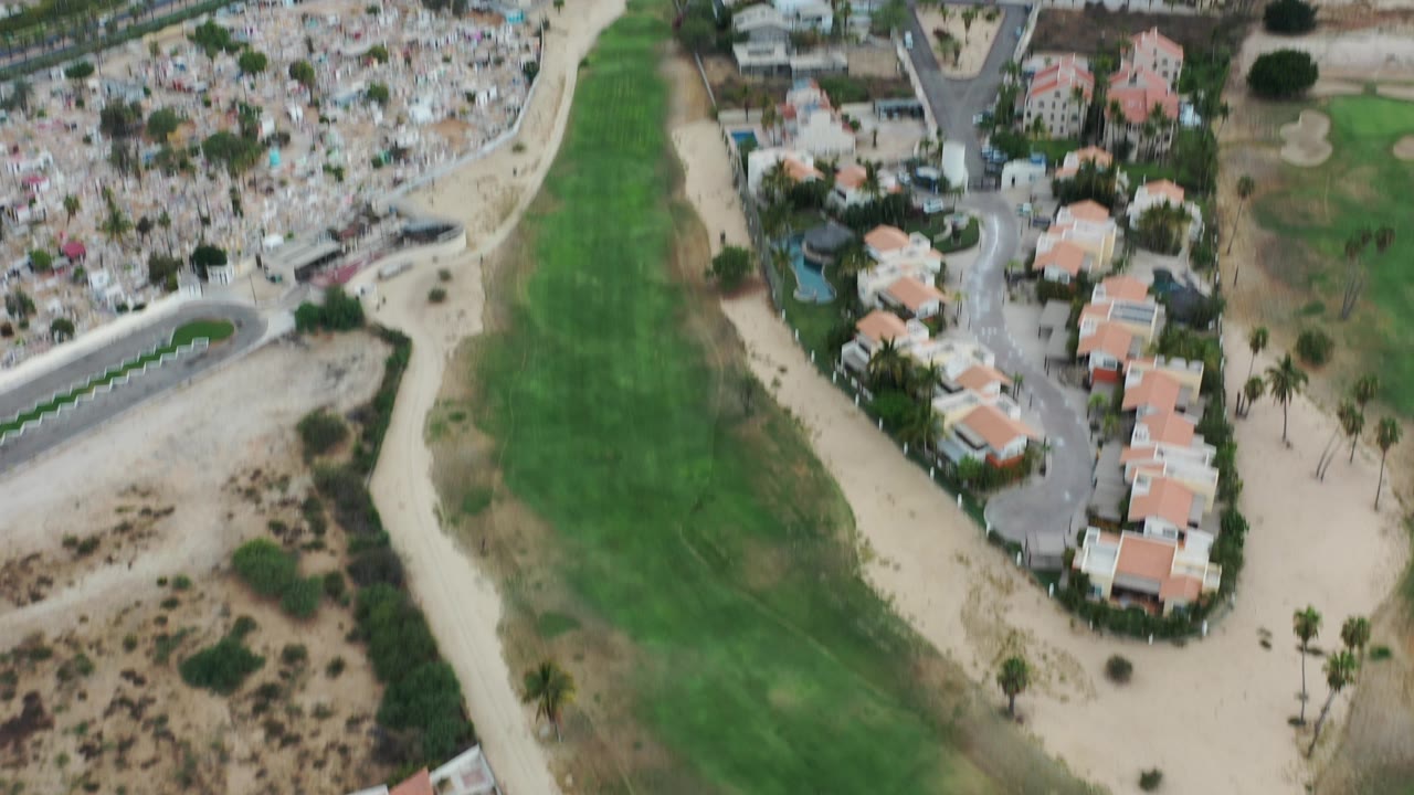 High aerial view over green fairway, roads, homes, and dense cemetery-like grounds.