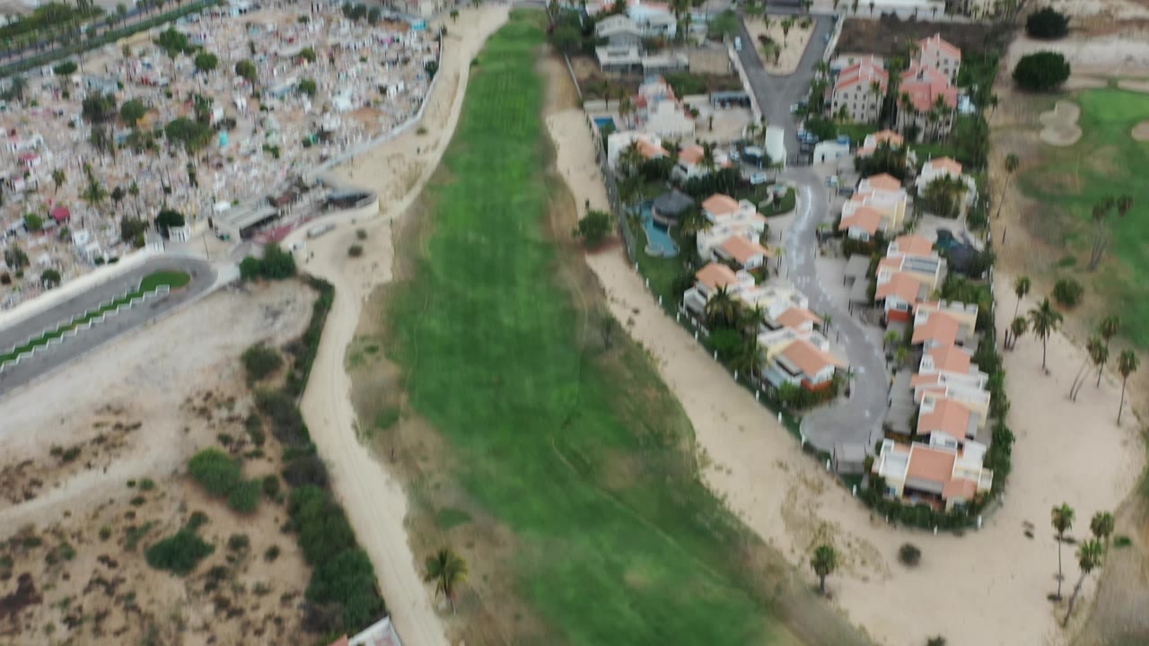 Los Cabos aerial view with a long green fairway centered between sandy lots, residential buildings, curving roads, and palm trees, with no visible people.