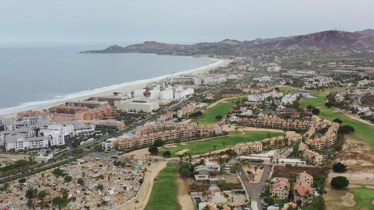 Aerial view over Los Cabos, Mexico with a curving beach and ocean on the left, golf greens and white buildings below, and dry hills in the distance, no visible people.