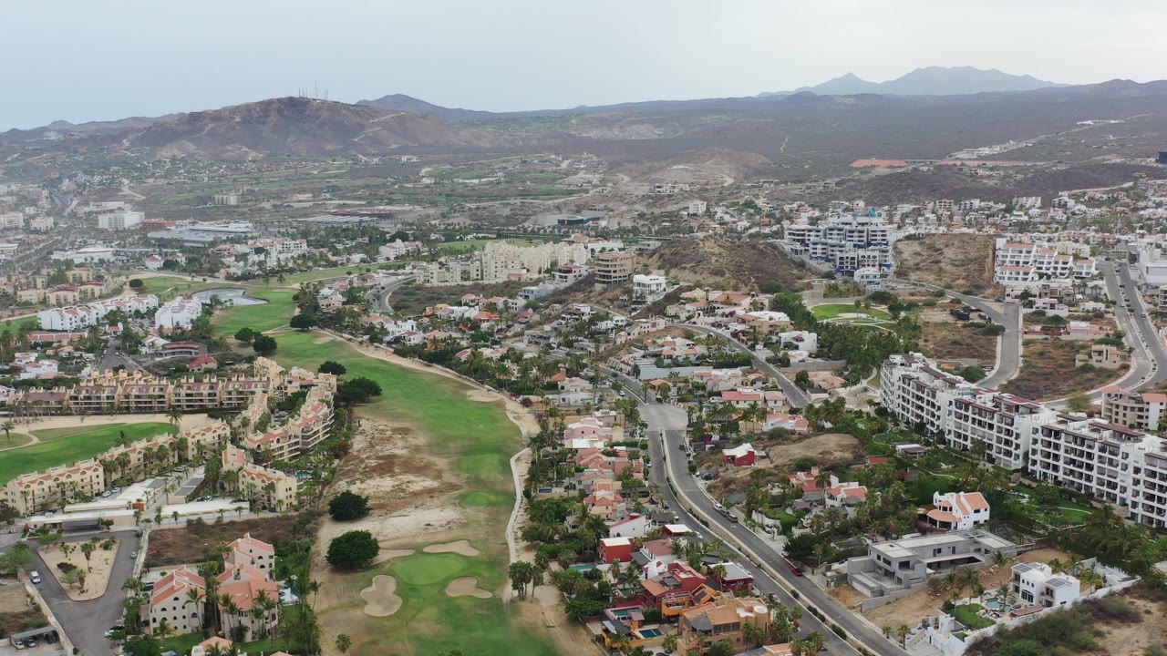 A slightly shifted Los Cabos aerial view centers the road corridor, apartment buildings, green fairways, dry hillside terrain, and no visible people.