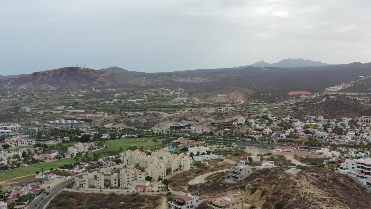 Wide aerial frame over Los Cabos, Mexico, showing residential buildings, dry hills, green fairway areas, distant mountains, overcast sky, and no visible people.
