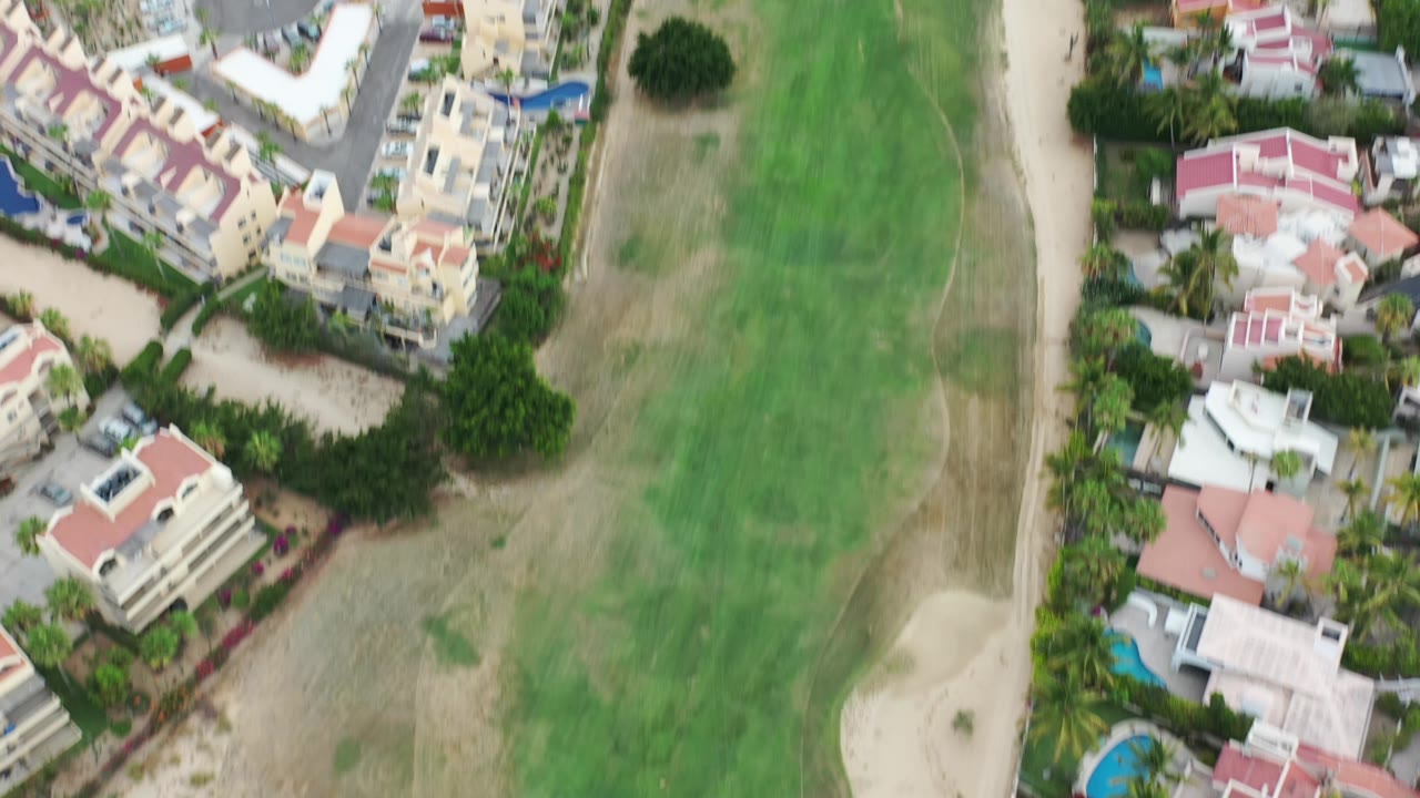 Overhead Los Cabos view with clustered houses, pale roofs, palms, a broad green fairway, and no visible people.