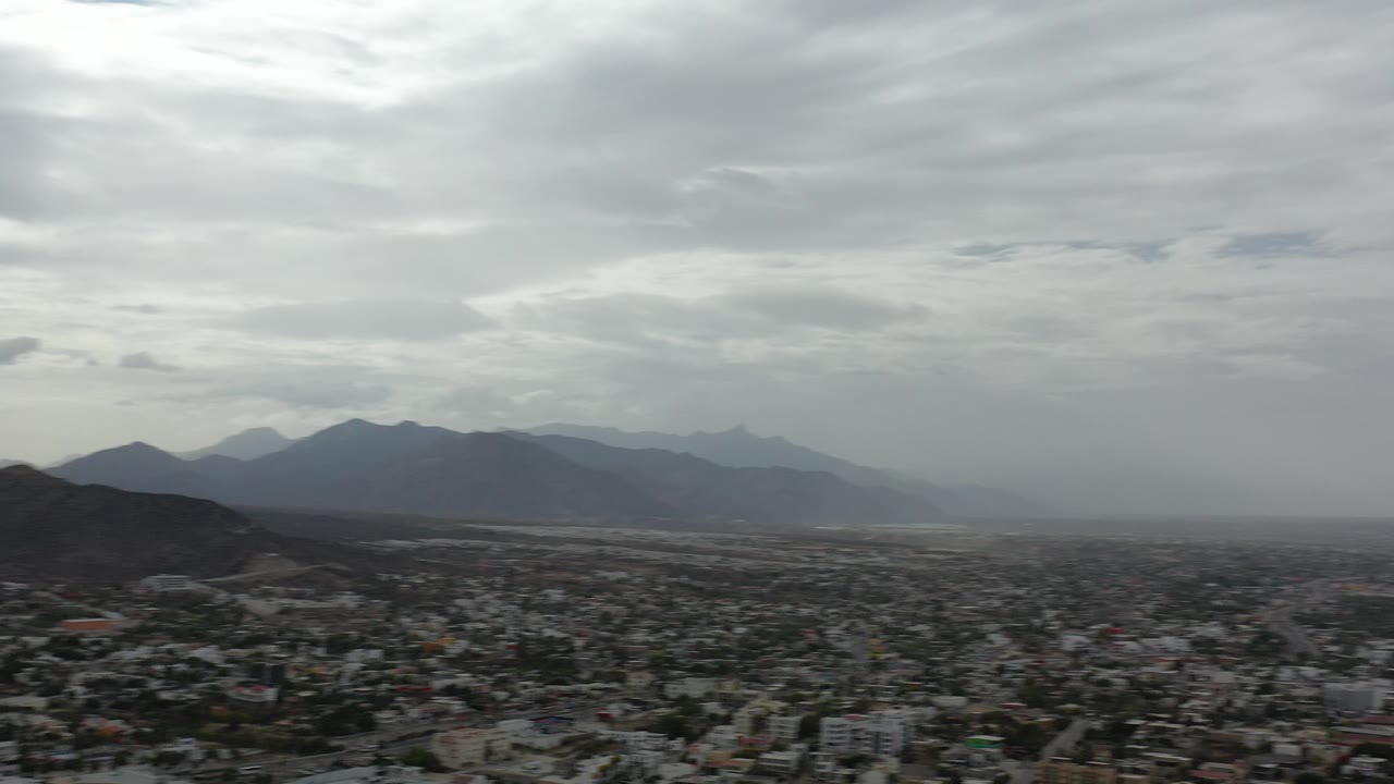 Wide aerial view over Los Cabos, Mexico, showing dense low-rise buildings, dark hills, distant mountains, gray cloud cover, and no visible people.