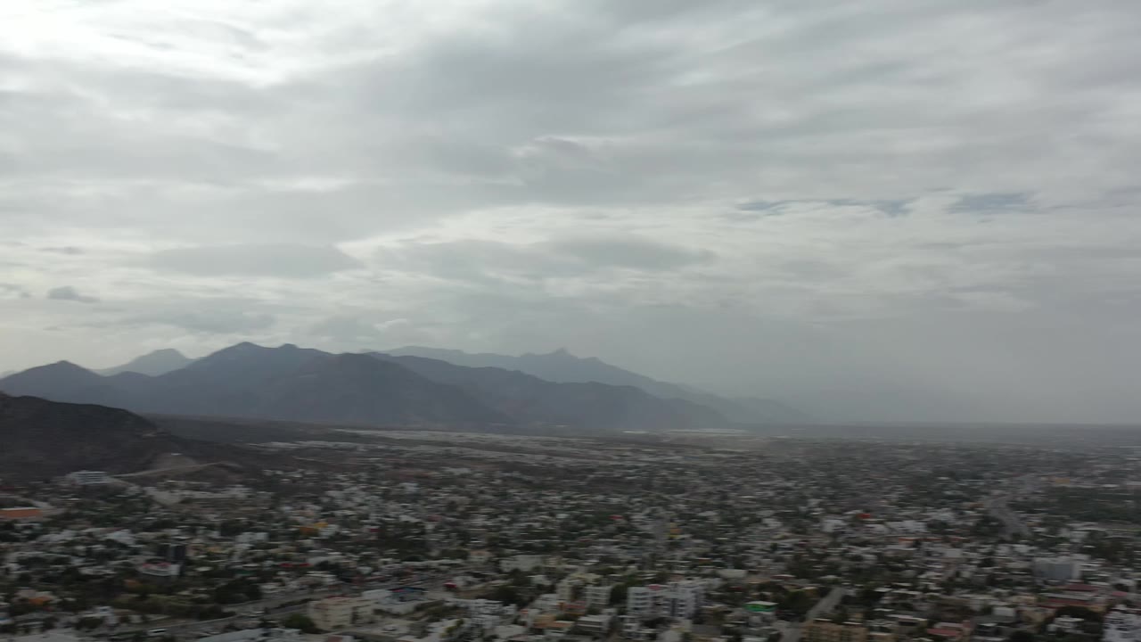 Aerial panorama in Los Cabos, Mexico, with the town spread across the lower frame, a low hill near center, layered mountains beyond, and no visible people.