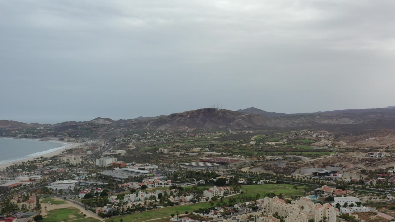 Wide aerial view over Los Cabos, Mexico with a curved bay at left, clustered buildings, green fairways, dry hills in the distance, and no visible people.