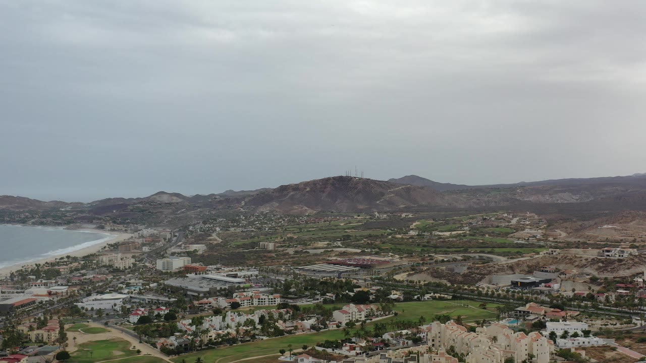 Wide drone view of coastal buildings, golf course, beach, and hills.