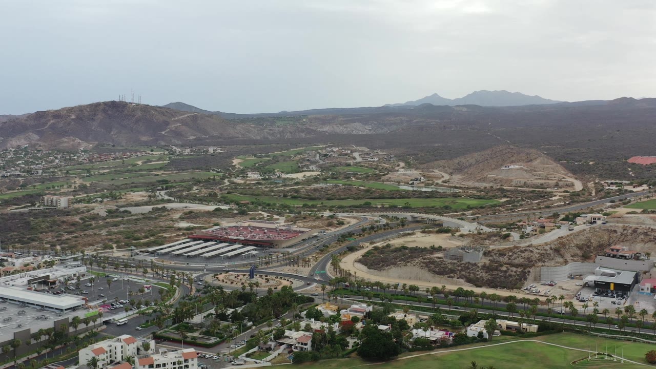 Wide aerial view of roads, buildings, green areas, and dry hills under cloudy sky