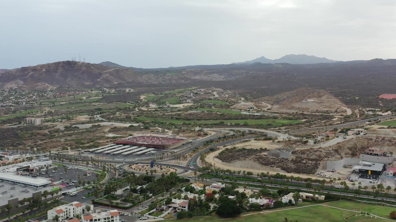 Aerial frame in Los Cabos with a road junction and parking areas in the foreground, green parcels through the valley, brown hills beyond, and no visible people.