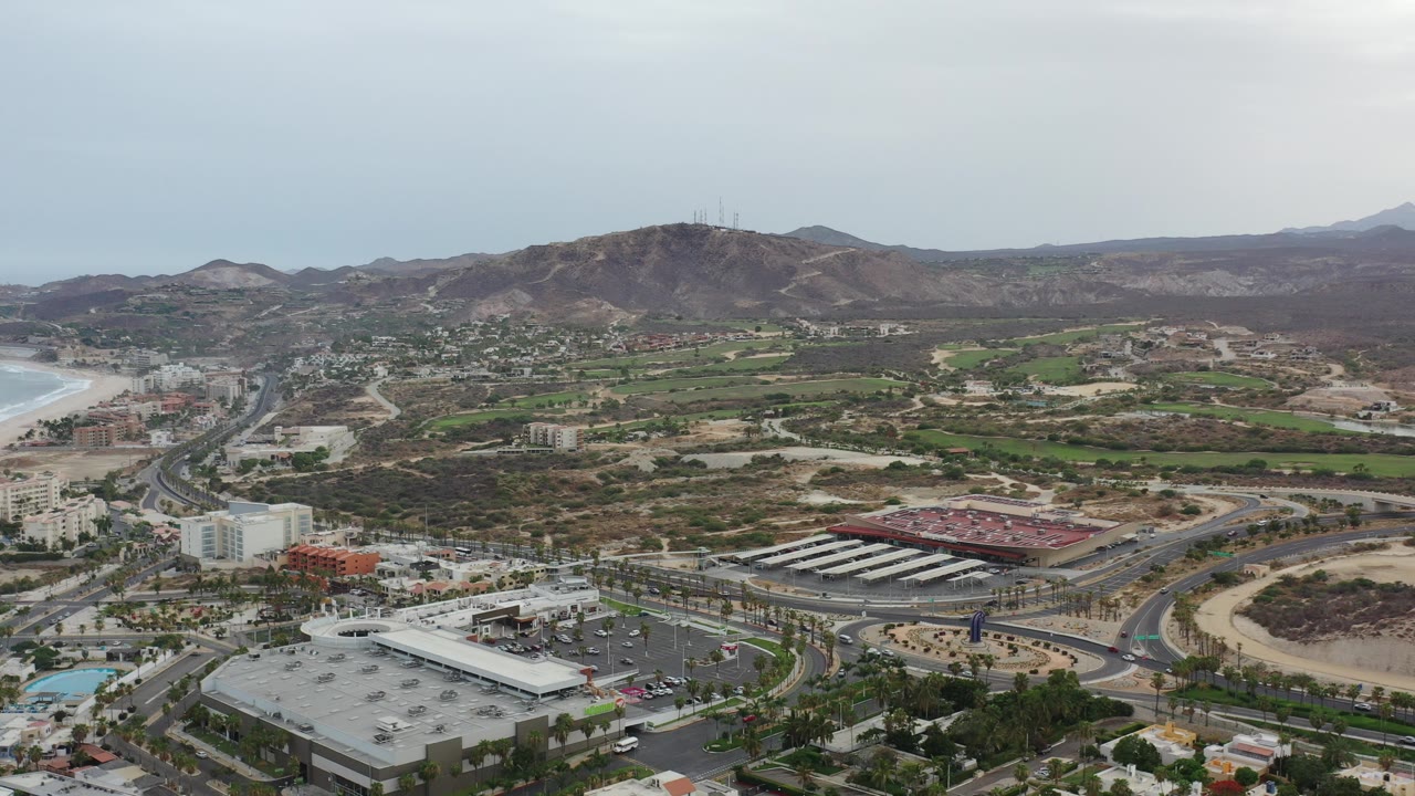 Opening aerial frame over Los Cabos, Mexico, showing a curving shoreline, city roads, low-rise buildings, green patches, dry hills, and no visible people.