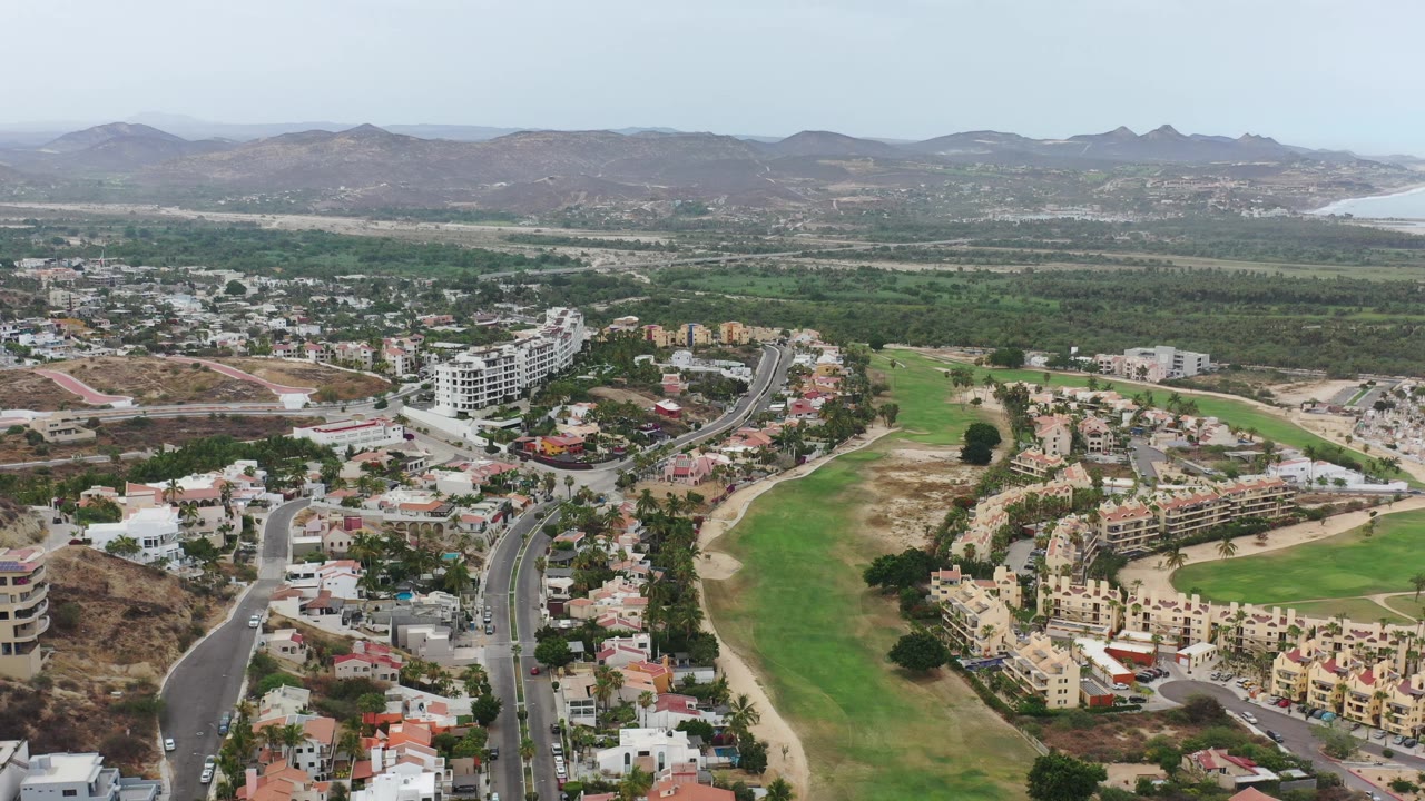 Aerial view of roads, homes, green fairway, mountains, and distant coast.