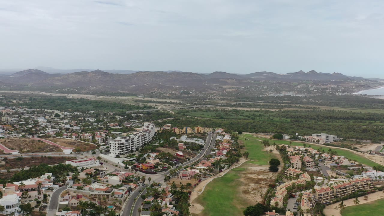 Wide aerial view of Los Cabos neighborhoods, green terrain, hills, and distant coastline.